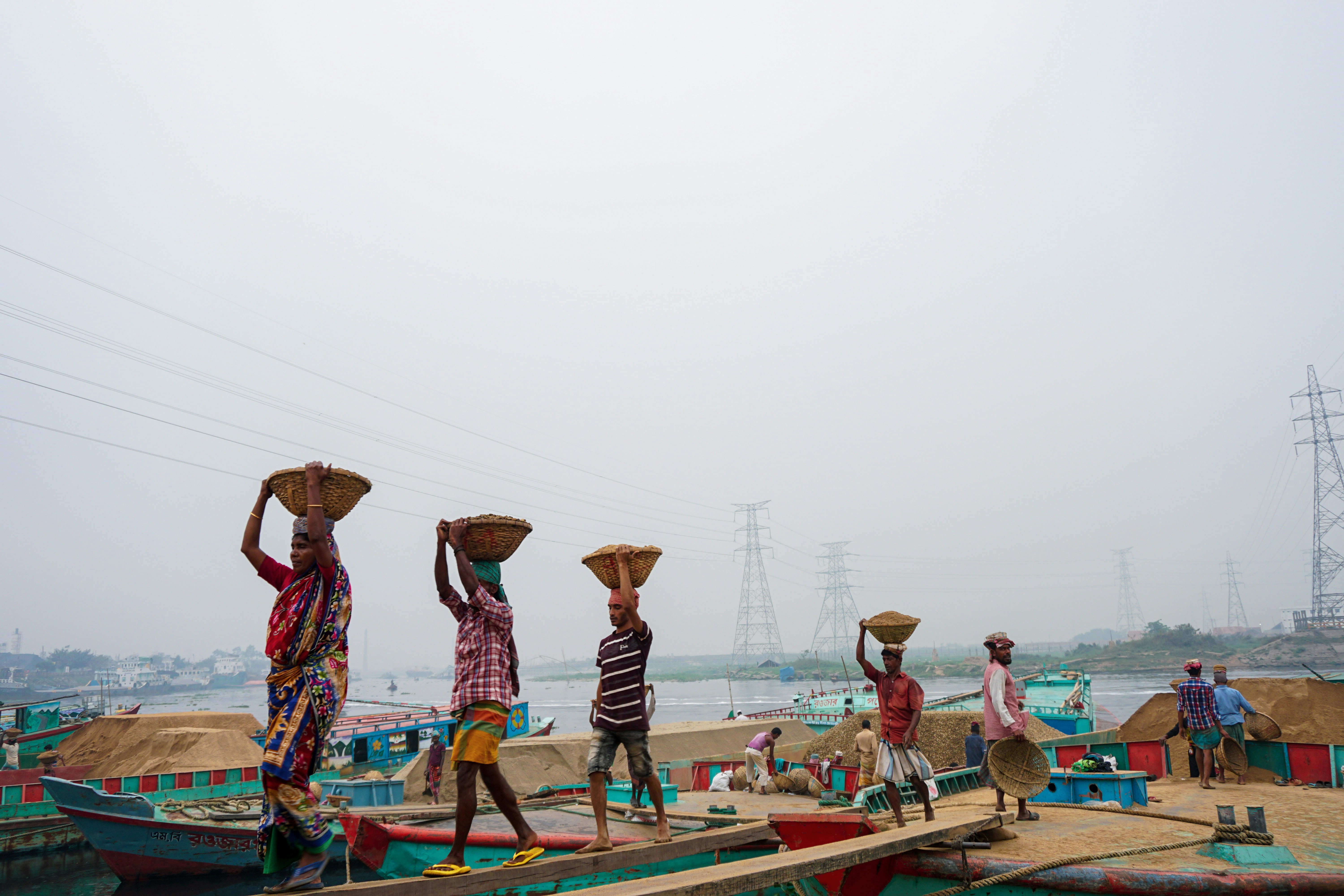 Workers carrying grain baskets along the riverfront in Bangladesh, illustrating daily labor and food supply challenges